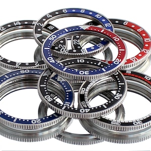 Op de afbeelding: Een close-up van een groep van acht horlogebezelen. De bezelen zijn gemaakt van metaal en hebben verschillende kleuren, waaronder zwart, blauw, rood en wit. De bezelen zijn genummerd van 0 tot 60.