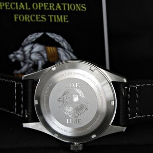 May include: A stainless steel watch with a black leather strap. The watch back features the text "S.O.F. TIME" and an emblem. The watch is set against a dark background with the text "SPECIAL OPERATIONS FORCES TIME". The strap has white stitching.