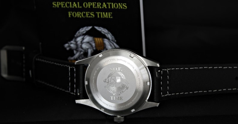 May include: A stainless steel watch with a black leather strap. The watch back features the text "S.O.F. TIME" and an emblem. The watch is set against a dark background with the text "SPECIAL OPERATIONS FORCES TIME". The strap has white stitching.
