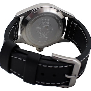 May include: A close-up view of a stainless steel watch with a black leather strap. The watch features a silver case and a black strap with white stitching. The watch's back has engraved text and a logo. The strap is fastened with a silver buckle.