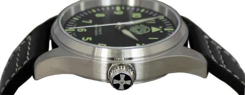 May include: A stainless steel wristwatch with a black face and luminous green numerals. The watch has a black leather strap with white stitching. The watch crown features a decorative design. The watch is an automatic.