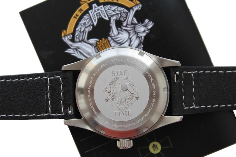 May include: A close-up of a stainless steel watch with a black leather strap and white stitching. The watch face features the text "S.O.F." and "TIME", with an emblem of two animals. It is a special edition for the Ukrainian Armed Forces.