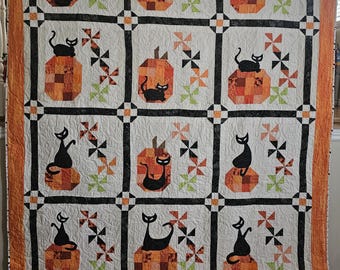 May include: A quilt featuring a grid of nine blocks, each displaying a black cat silhouette perched on a patchwork pumpkin. The design includes small pinwheel motifs in orange, black, and green, separated by black sashing and an orange border.