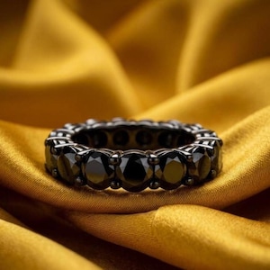 May include: A black eternity ring featuring numerous round-cut black gemstones. The ring is set in a dark metal and is displayed on a golden, textured fabric. The gemstones are evenly spaced around the band, forming a continuous circle of black.
