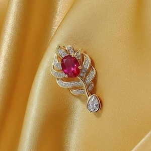May include: A gold-toned feather brooch with a large oval ruby gemstone at the top and a pear-shaped clear gemstone at the bottom. The feather design is embellished with small, sparkling clear gemstones.