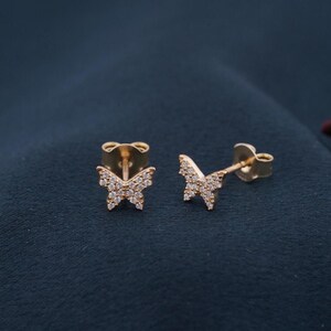May include: A pair of gold-colored butterfly stud earrings. Each earring features a butterfly design encrusted with small, clear gemstones. The earrings are set against a dark blue background.
