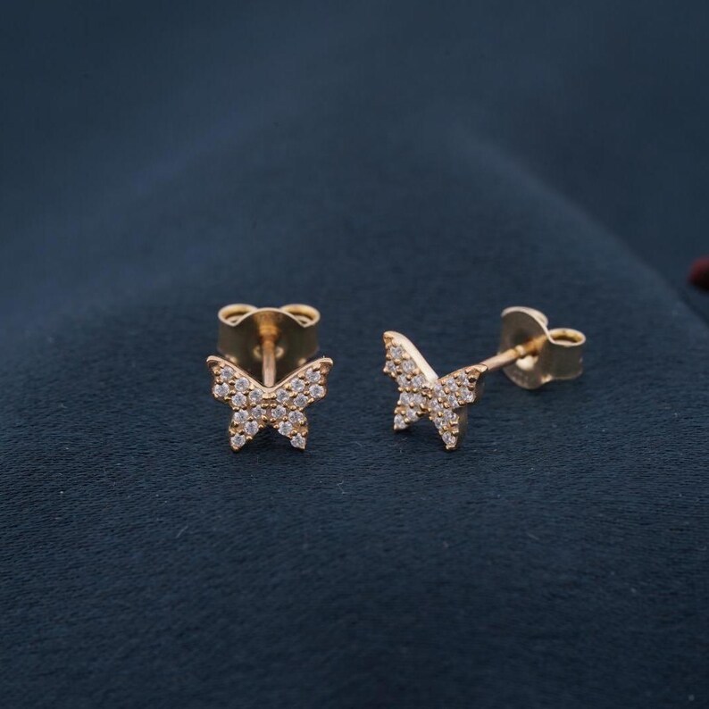 May include: A pair of gold-colored butterfly stud earrings. Each earring features a butterfly design encrusted with small, clear gemstones. The earrings are set against a dark blue background.