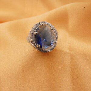 Könnte beinhalten: Ein silberner Ring mit einem großen, oval geschliffenen, blau-klaren Edelstein. Der Edelstein ist von kleineren, klaren Steinen umgeben. Der Ring liegt auf goldfarbenem Stoff.