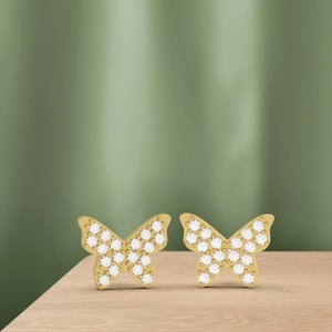 May include: A pair of gold-colored butterfly stud earrings. Each earring features a butterfly shape with a surface covered in small, sparkling white stones. The earrings are displayed on a light brown wooden surface against a green background.