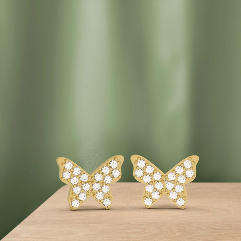 May include: A pair of gold-colored butterfly stud earrings. Each earring features a butterfly shape with a surface covered in small, sparkling white stones. The earrings are displayed on a light brown wooden surface against a green background.