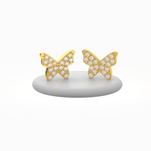 May include: A pair of gold-colored butterfly stud earrings. Each earring features a butterfly shape encrusted with small, clear gemstones. The earrings are displayed on a white, rounded surface against a white background.