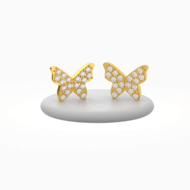 May include: A pair of gold-colored butterfly stud earrings. Each earring features a butterfly shape encrusted with small, clear gemstones. The earrings are displayed on a white, rounded surface against a white background.