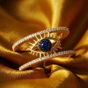 Könnte beinhalten: Ein goldener Ring mit einem augenförmigen Mittelstück, das einen dunkelblauen Stein umgeben von kleinen schwarzen Steinen zeigt. Das Augenmotiv hat strahlende goldene Strahlen und wird von zwei mit kleinen weißen Steinen besetzten Bändern flankiert.