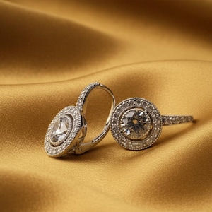 May include: Silver earrings featuring a central clear stone encircled by smaller stones. The earrings are set on a golden satin backdrop. The design is elegant and the overall aesthetic is luxurious.