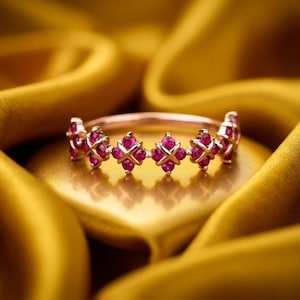 May include: A rose gold ring with a repeating pattern of pink gemstones. The gemstones are set in a floral or cross-like design. The ring is displayed on a golden, silky fabric.