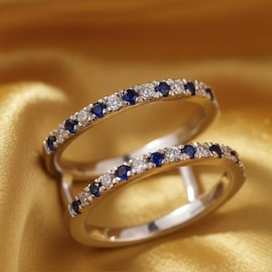May include: Two silver rings with a row of small, round, blue gemstones alternating with clear gemstones. The rings are set against a gold-colored, textured background.