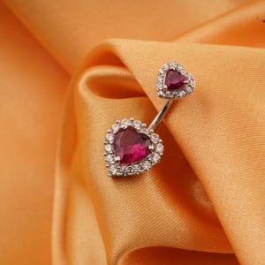 Może przedstawiać: Srebrny kolczyk do pępka z dwoma kamieniami szlachetnymi w kształcie serca. Większe serce jest w kolorze głębokiej czerwieni, otoczone aureolą z małych, przezroczystych kamieni. Mniejsze serce u góry ma również czerwony kamień i aureolę z przezroczystych kamieni.