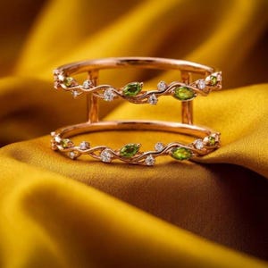 May include: A double-band ring crafted from rose-colored metal, featuring a vine-like design adorned with marquise-cut green peridot stones and small, round, clear white accent stones set along the delicate, intertwining metal branches.