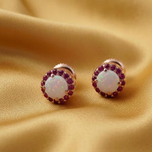 May include: A pair of stud earrings featuring a central white opal stone surrounded by a halo of small, round, deep red gemstones. The earrings are set in a rose gold metal and rest on a golden, textured surface.
