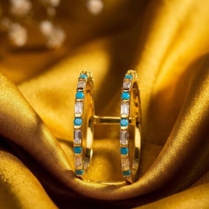 May include: A gold open-style ring featuring two parallel bands connected by a bridge. Each band is set with alternating clear baguette-cut stones and small round turquoise-colored stones arranged in a repeating pattern along the outer edge.