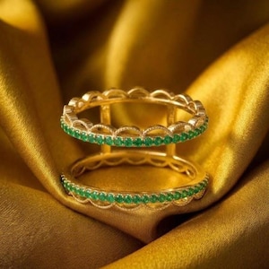 May include: A double-band gold ring featuring two parallel bands connected by vertical gold bars. Each band is encrusted with a continuous row of small, round, vibrant green gemstones set in a scalloped, decorative metal frame design.