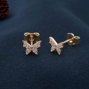 May include: A pair of gold-colored butterfly stud earrings. Each butterfly is encrusted with small, clear gemstones. The earrings are displayed on a dark blue surface, showcasing their delicate design and sparkle.