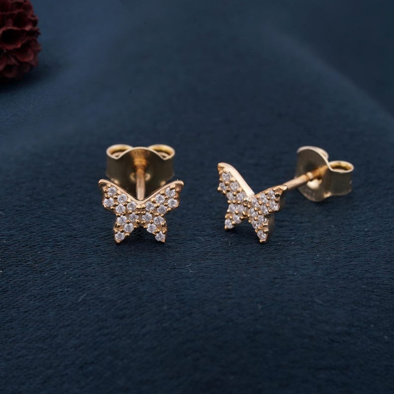 May include: A pair of gold-colored butterfly stud earrings. Each butterfly is encrusted with small, clear gemstones. The earrings are displayed on a dark blue surface, showcasing their delicate design and sparkle.