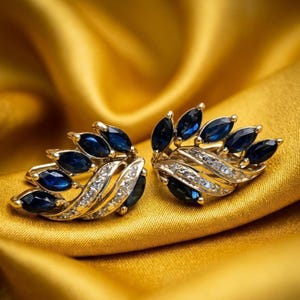 Könnte beinhalten: Ein Paar goldfarbene Ohrringe mit einem blattartigen Design. Jeder Ohrring hat dunkelblaue, marquiseförmige Edelsteine und kleine, klare Edelsteine. Die Ohrringe werden auf einem goldenen, strukturierten Stoff präsentiert.