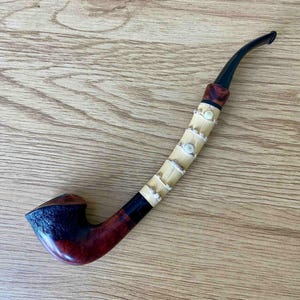 May include: A tobacco pipe featuring a dark reddish-brown wooden bowl with a textured finish, a light-colored bamboo shank accented with silver-toned metal rings and small circular inlays, and a curved black acrylic stem at the end.