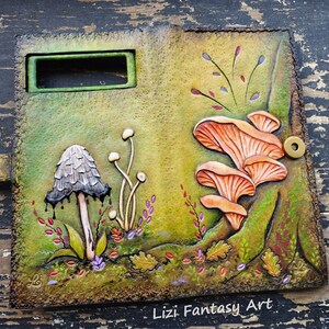 May include: A leather journal cover with a hand-tooled design featuring mushrooms, leaves, and flowers. The cover is green, brown, and orange with a small brass button. The text "Lizzi Fantasy Art" is embossed on the bottom of the cover.