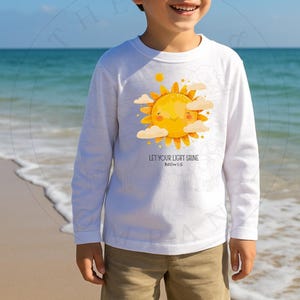 May include: White long-sleeve shirt featuring a smiling yellow sun graphic with clouds. The shirt has the text "LET YOUR LIGHT SHINE" and "Matthew 5:16". The shirt is worn with khaki shorts.