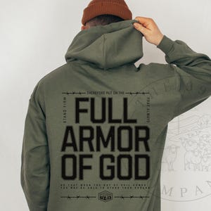 May include: Olive green hoodie with the text "FULL ARMOR OF GOD" in bold, black letters. The design includes additional text and a graphic of sheep. The hoodie has a drawstring hood and long sleeves.