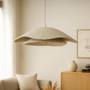 Pode incluir: Uma luminária pendente bege em camadas pende do teto. A luz tem uma forma texturizada e orgânica, que se assemelha a pétalas sobrepostas. A luminária está suspensa acima de uma sala de estar com um sofá, obras de arte e um armário de madeira.