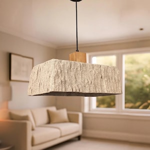 Puede incluir: Una lámpara colgante rectangular con una pantalla texturizada, similar a la piedra. La pantalla es de color beige claro y está suspendida del techo por un cable negro y un bloque de madera. La lámpara está colgada en un salón.