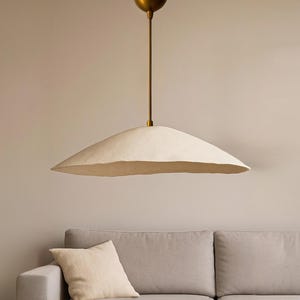May include: A cream-colored, saucer-shaped pendant light fixture with a brass-colored stem and ceiling mount. The light hangs above a gray sofa with a matching pillow. The fixture has a textured, paper-like shade.