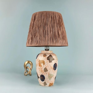 May include: A table lamp with a brown lampshade and a patterned base. The base has a speckled design with orange, brown, and blue accents. The lamp is topped with a brown, pleated lampshade. The lamp is approximately 50 cm tall.