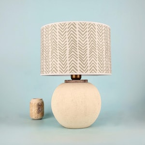 May include: A cream-coloured table lamp with a round base and a patterned lampshade. The lampshade has a repeating chevron design in light grey. A small decorative vase with a geometric pattern sits beside the lamp.
