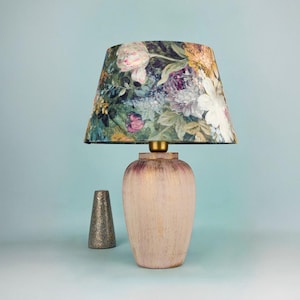 May include: A table lamp featuring a floral lampshade, with a light pink, distressed base. A small, silver-coloured, cone-shaped object is positioned to the left of the lamp. The lamp is set against a light blue backdrop.