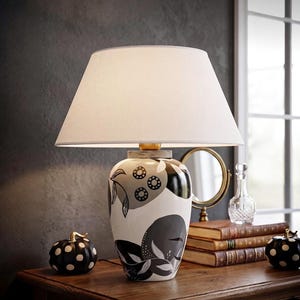 May include: A table lamp with a white tapered fabric shade and a ceramic base featuring a black and white abstract botanical pattern. The lamp sits on a wooden surface alongside two black pumpkins with white polka dots and a stack of books.