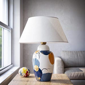 Può includere: Una lampada da tavolo con paralume conico in tessuto bianco e base in ceramica. La base è bianca con forme astratte dipinte in blu navy, azzurro e giallo senape, arricchite da sottili linee nere e dettagli circolari color oro.