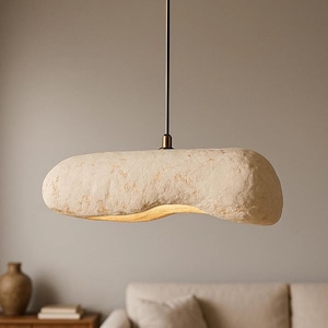 K&ouml;nnte beinhalten: Eine cremefarbene, l&auml;ngliche Pendelleuchte mit strukturierter Oberfl&auml;che. Das Licht h&auml;ngt an einer bronzefarbenen Stange und einem kreisf&ouml;rmigen Baldachin. Die Leuchte h&auml;ngt &uuml;ber einem Wohnzimmer.