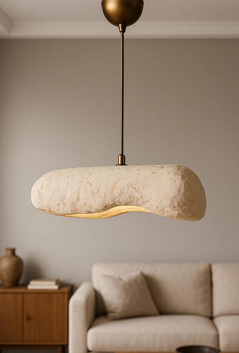 K&ouml;nnte beinhalten: Eine cremefarbene, l&auml;ngliche Pendelleuchte mit strukturierter Oberfl&auml;che. Das Licht h&auml;ngt an einer bronzefarbenen Stange und einem kreisf&ouml;rmigen Baldachin. Die Leuchte h&auml;ngt &uuml;ber einem Wohnzimmer.