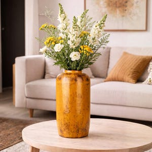 Op de afbeelding: Een hoge, cilindervormige, gele vaas met een verweerde afwerking bevat een boeket witte en gele bloemen met groen blad. De vaas staat op een ronde, lichtgekleurde houten tafel. Op de achtergrond staat een beige bank.