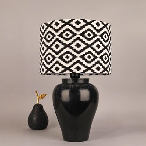 May include: A black table lamp with a patterned lampshade. The lampshade has a black and white geometric design. A black pear-shaped decorative object with a gold leaf is next to the lamp.