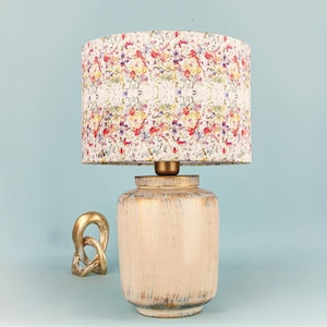 May include: A cream-coloured table lamp with a floral patterned lampshade. The lamp base has a distressed finish with gold accents. A gold-coloured decorative knot is to the left of the lamp.
