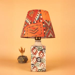 May include: A decorative table lamp with a patterned orange lampshade and a white base adorned with geometric designs in red, green, and black. The lamp has a brass-coloured neck and sits on a light orange surface.