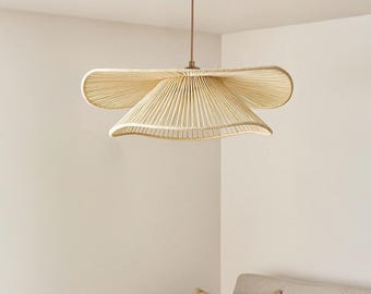May include: A beige woven pendant light fixture with a unique, layered design. The light hangs from a bronze-colored ceiling mount and rod. The fixture's shape resembles a stylized flower or hat, adding a touch of natural elegance to the room.