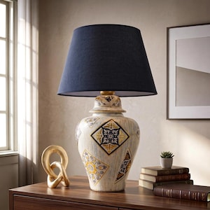 Op de afbeelding: Een tafellamp met een donkerblauwe stoffen kap in trommelvorm en een keramische voet met een verweerde witte afwerking. De voet is versierd met gouden en donkerblauwe geometrische ruitpatronen en bloemmotieven, afgewerkt met een gouden hals.