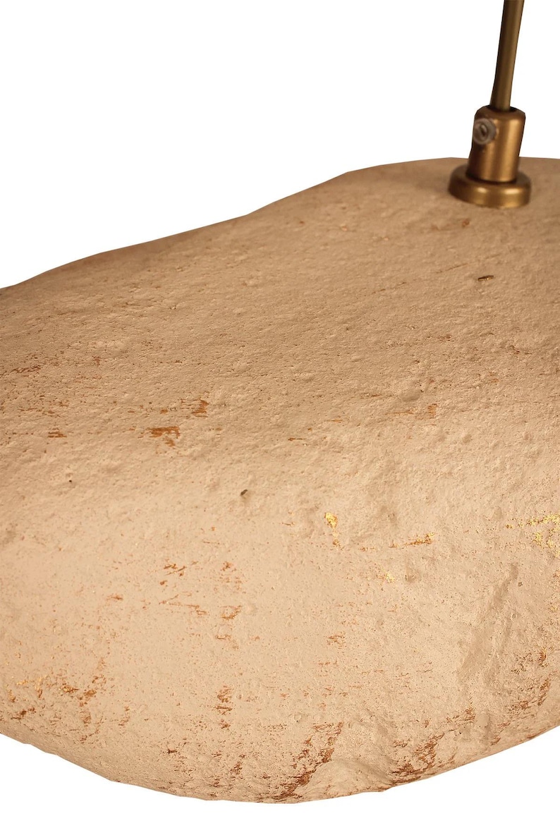 K&ouml;nnte beinhalten: Eine beige, organisch geformte Pendelleuchte. Die Leuchte hat eine strukturierte Oberfl&auml;che mit dezenten Goldakzenten. Ein messingfarbener Stiel und eine Halterung verbinden sich mit der Decke.
