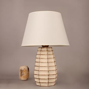 May include: Cream-colored table lamp with a white lampshade. The lamp base has a textured, grid-like design with gold accents. A small, decorative vase with a geometric pattern sits beside the lamp. Home decor.